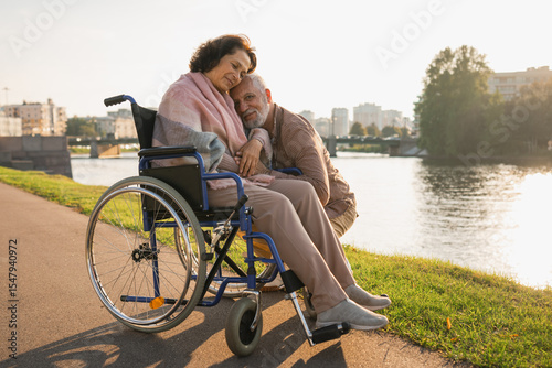 Senior woman in wheelchair walking with caregiver old man on road. Elderly family couple man supporting taking care of paralyzed woman in chair for people with disability outdoor. Rehabilitation