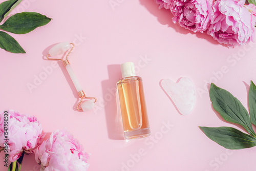 Cosmetic bottle, face roller, gua sha stone and pink peonies on pastel pink background, skincare concept. Top view, flat lay