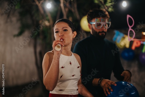 Two individuals at a lively party, one blowing a noisemaker, surrounded by balloons and festive decor, creating a joyful, celebratory atmosphere.