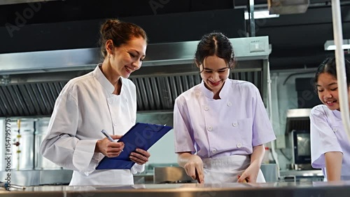 Female chef teacher teaches two students to cook in class. A cooking test is given to the teenage girls to cook spaghetti. The teacher holds a file to give scores in the cooking class.