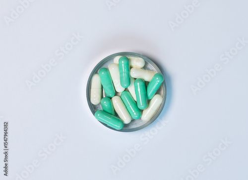Close-up top-view of green and white capsule pills in a metal container on white background. Lay flat composition shows pharmaceutical use, healthcare concepts, and clean, medical product.