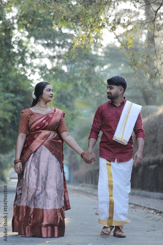 A beautiful South Indian couple dressed in elegant traditional attire walking hand in hand along a serene tree-lined path. The woman is wearing a silk saree with jewelry and jasmine flowers in her hai A beautiful South Indian couple dressed in elegant traditional attire walking hand in hand along a serene tree-lined path. The woman is wearing a silk saree with jewelry and jasmine flowers in her hai