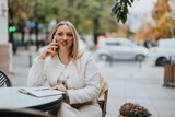 Businesswoman in an elegant outfit is smiling and talking on a phone outdoors. She's seated at a table with a notebook, enjoying a pleasant conversation during a modern city day.