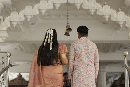An Indian couple performs the sacred Abhishekam ritual to a Shiva Lingam in a Hindu temple. The woman is dressed in an elegant silk saree with jasmine flowers in her hair, and the man wears traditiona