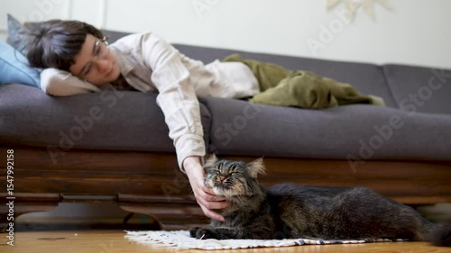 Relaxed woman lounging on sofa strokes cat lying on floor mat nearby, enjoying calm weekend together. Pet bonding, indoor leisure, animal pet companionship, slow living, domestic mutual comfort
