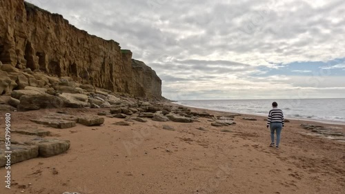 A women walking along a Jurassic Coast beach in Dorset. The camera moves sideways to the right as the female walks away along the beach wide the sea on the right and the cliffs left.