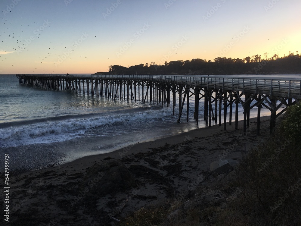 Obraz premium A picturesque wooden pier stretches into the Pacific Ocean at San Simeon, California, bathed in the warm glow of a sunset, inviting tranquility.
