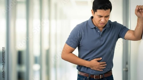 An asian man experiencing abdominal pain, leaning against a wall and holding his stomach in discomfort.