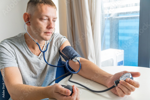 Man checking blood pressure manually with sphygmomanometer and stethoscope while sitting at home. Managing hypertension, heart health and general wellness. Regular self health monitoring