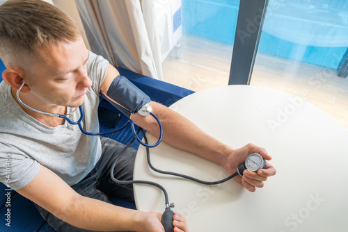 Man using manual sphygmomanometer and stethoscope to check his own blood pressure while sitting at home. Managing hypertension, heart health and general wellness. Regular self health monitoring