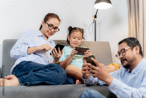 Happy Asian parents and child enjoying screen time together in living room, each on their own smartphone.