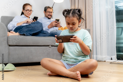Happy Asian parents and child enjoying screen time together in living room, each on their own smartphone.