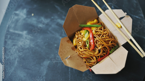A high-quality food photograph of Chinese lo mein noodles served in a brown paper takeout container with wooden chopsticks