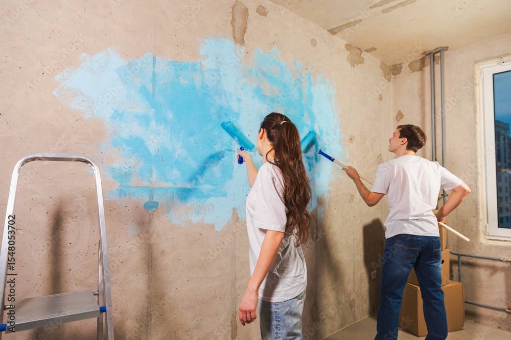 Obraz premium Couple in new home during repair works painting wall together. Happy family holding paint roller painting wall with blue color paint in new house. Home renovation DIY renew home concept