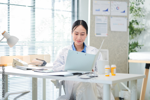 Portrait of a confident Asian female doctor smiling while working at her desk in a clinic, typing on her laptop and reviewing medical reports. Modern healthcare professional indoors with document file