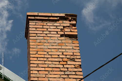 Chimney displays significant wear and damage with cracks against a backdrop of a clear blue sky, indicating possible structural concerns