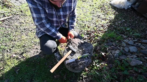 A skilled artisan carefully chisels a piece of stone while kneeling in a sunlit garden. Surrounding leaves reflect the vibrant colors of autumn, creating a serene atmosphere.