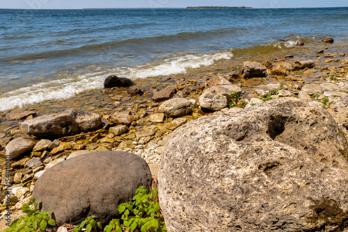 The large pitted boulder captivates the viewer as the water of Green Bay wash ashore at Peninsula State Park, Fish Creek, Wisconsin in mid-June, as a distant islands rises from before the horizon