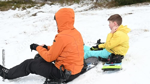 A man and a boy, bundled in winter gear, pause their snowboarding adventure. They sit in the snow, enjoying a moment of rest. The scene exudes a sense of winter fun and family time.