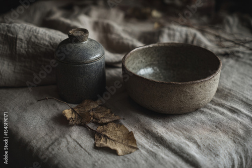 Minimal still life with hot water bottles, dried leaves, and ceramic bowl on linen cloth