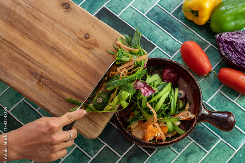 Detailed view of discarded vegetable skins and trimmings on a rustic kitchen surface. Onion layers, carrot ribbons, and zucchini strips in a natural, eco-conscious kitchen with vintage green tiles.
