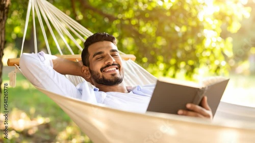 Smiling man relaxes in a hammock under trees He is reading a book enjoying the sunshine and the peacefulness of the day Peaceful leisure