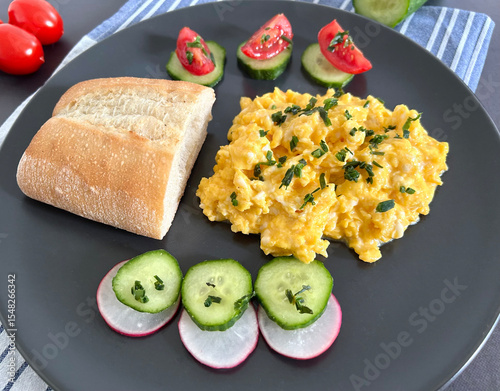 A fresh and nutritious breakfast plate featuring creamy scrambled eggs topped with chopped chives, served with ciabatta bread, cucumber and radish slices, and cherry tomatoes. Healthy Breakfast Plate.