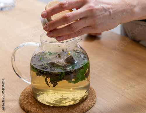 Woman opens wooden lid of the stylish transparent tea pot with just brewed green tea leaves, mint leaves and lemon slices in it. Beverage