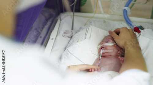 A premature baby lies in an incubator, hand held by an adult. The infant has medical equipment attached while resting on white linens.
