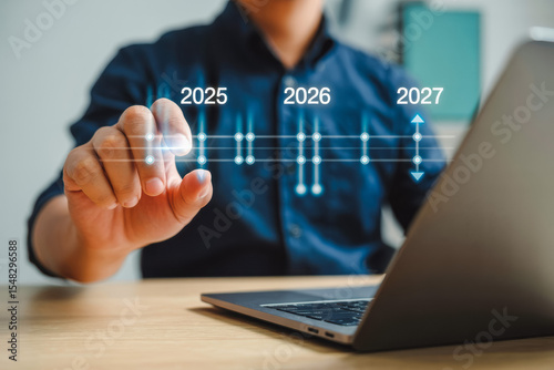 A person interacts with a futuristic timeline interface, highlighting years 2025, 2026 to 2027. They're seated at a desk with a laptop, symbolizing forward-thinking technology and innovation.