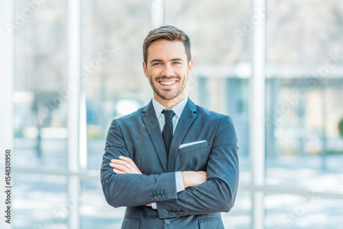 Professional businessman in a tailored suit stands confidently with arms crossed in a bright office setting. Smiling, with natural light pouring in, enhancing a crisp, modern ambiance.