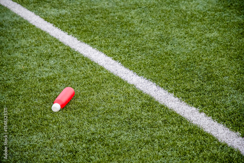red water bottle lies on the green artificial grass of a football field near the dividing white line, the concept of drinking during long workouts and replenishing fluids and electrolytes,
