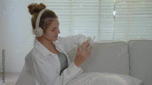 Cheerful woman enjoying relaxing lifestyle in her living room, using Facetime on her tablet while wearing headphones and lounging on cozy couch.