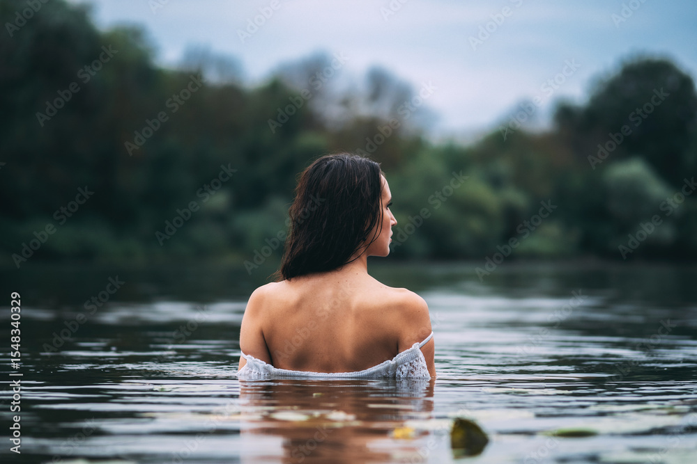 Beautiful long-haired brunette in the lake in a white dress. Portrait of a woman back view in the water in summer cloudy weather Beautiful long-haired brunette in the lake in a white dress. Portrait of a woman back view in the water in summer cloudy weather