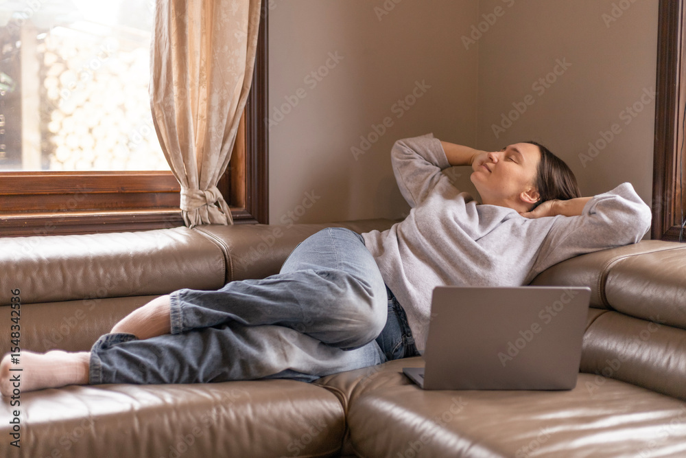 Fototapeta premium A woman stretches her neck while lying on the couch in front of her laptop