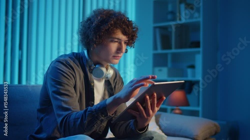Young Man Sits on Sofa with Tablet in Hands, Working with Concentration. Calm and Efficient Evening in a Flexible Tech-Driven Workflow. Focus Mode, Digital Productivity, Self-Management.