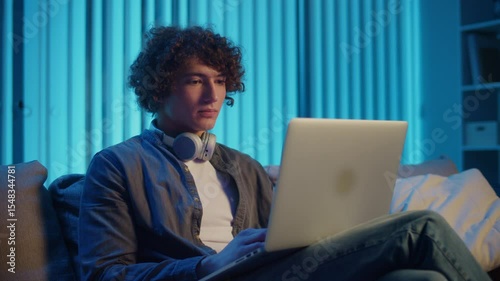 Young Man Sits on Sofa in His Room at Night, Studying or Working on Laptop. Evening Routine of a Student in a Tech-Driven World. Remote Learning, Digital Productivity, Mental Focus.