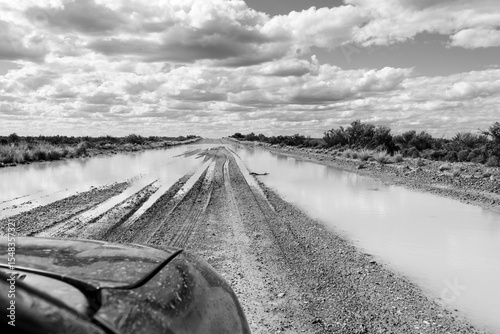 Black and white image showing the front of a car facing a large mud puddle on a dirt road
