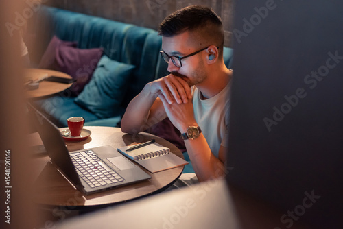 Focused young man wearing glasses and wireless earbuds, watching a webinar, having an online course on a laptop in a modern interior. 