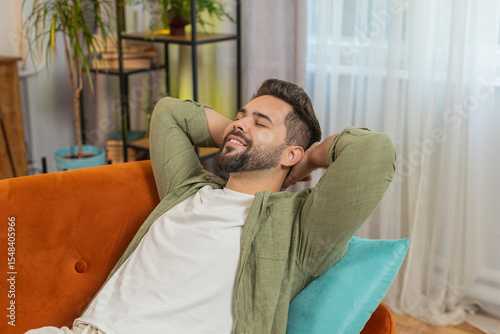 Tired Caucasian handsome man enjoy relaxing on home sofa in living room. Happy guy lying on couch resting napping after hard working day, closed his eyes, put his hand behind his head, stress free.