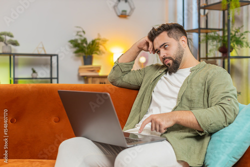 Overtired Caucasian young man with brown hair bored using laptop while sitting on sofa at home. Sleepy guy cannot concentrate on work and types lazily. Adult male is not interested in doing a project.