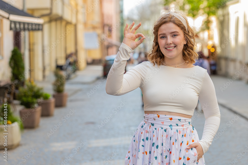 Naklejka premium Okay. Happy cheerful redhead young woman in long dress looking approvingly at camera showing ok gesture, positive like sign, approve something good, celebrate win outdoors. Girl in urban city street