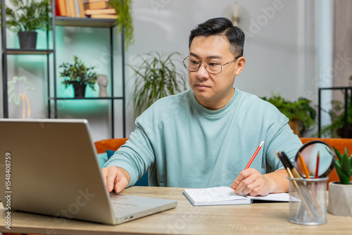 Asian man freelancer making notes on notebook at home, use laptop, reading information prepare business financial report. Guy accountant analyzing reports documents calculate taxes, studying education