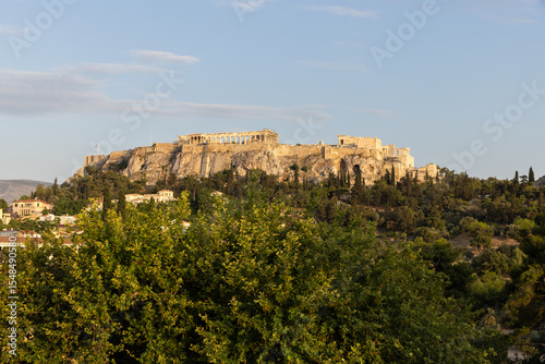 Landscape view of the Acropolis in Greece with blue sky and lush trees in the foreground. 