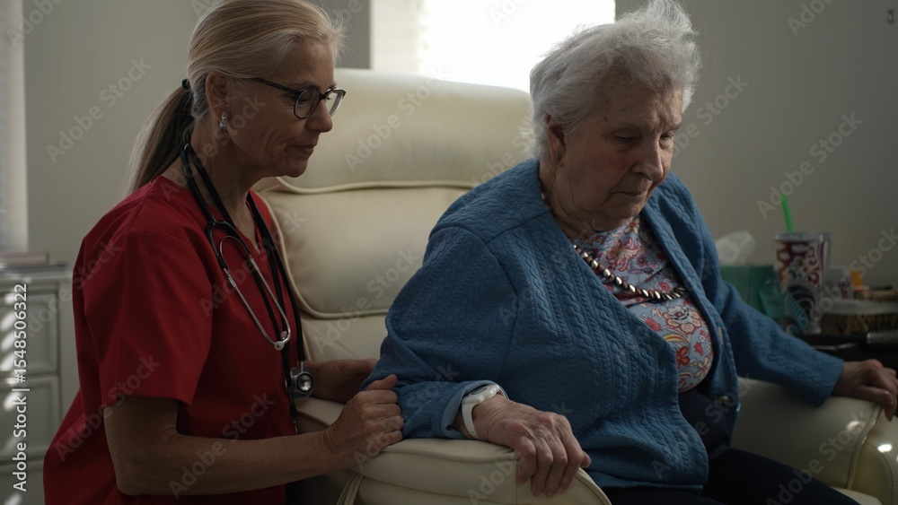 Obraz premium A caregiver in scrubs communicates with an elderly woman seated in a chair, fostering engagement and emotional support in a home-like healthcare environment.
