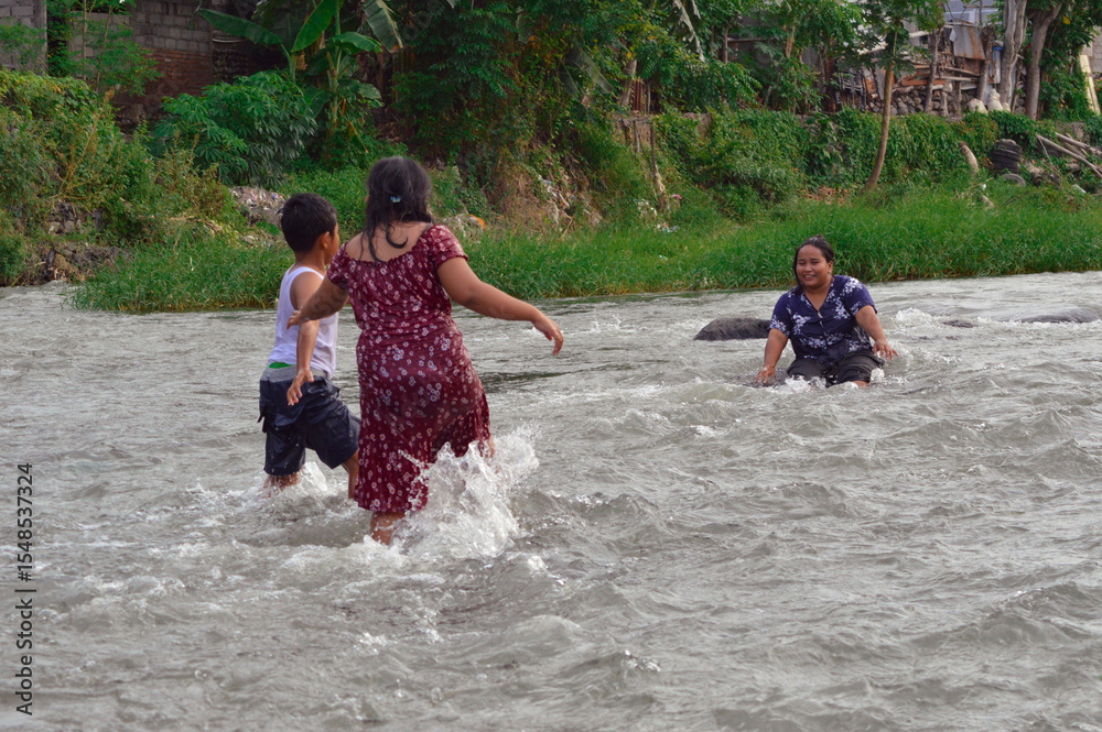 Fototapeta premium A mother sits with her legs outstretched in the stream, while two children play around her, walking and enjoying the refreshing water.