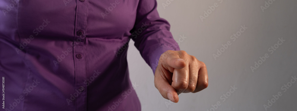 Fototapeta premium A close-up of a hand pointing forward, wearing a purple shirt with buttoned sleeve cuffs against a grey background. 