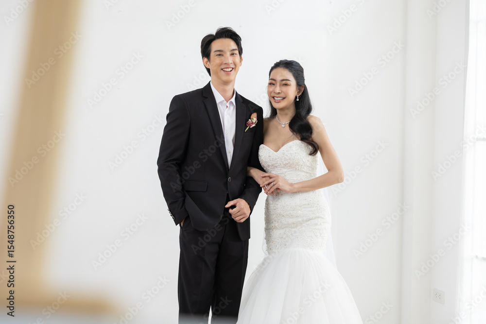 Asian Happy bride and groom smiling together in elegant wedding attire, standing in bright white room celebrating their romantic marriage ceremony Asian Happy bride and groom smiling together in elegant wedding attire, standing in bright white room celebrating their romantic marriage ceremony