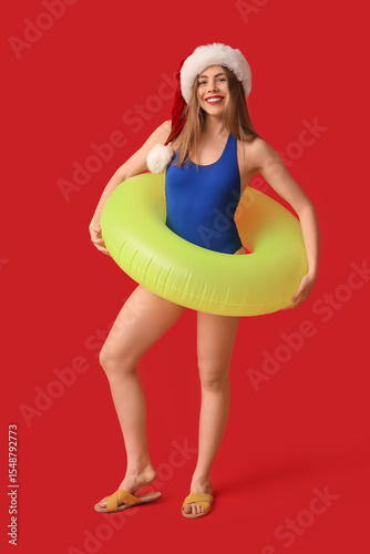 Happy young woman in swimsuit and Santa hat with inflatable ring on red backg...