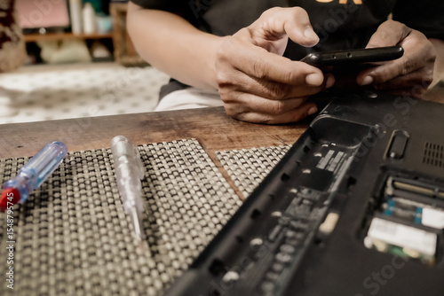 Male person searching for instructions from the internet using smartphone to repair a broken laptop at home. Do it yourself concept.